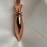 Close top-down view of a polished pure copper pendulum, long spindle shape, warm reflective finish on textured white fabric