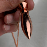Fingers holding the tip and chain loop, reflective scrying pendulum finish, classic copper dowsing pendulum feel